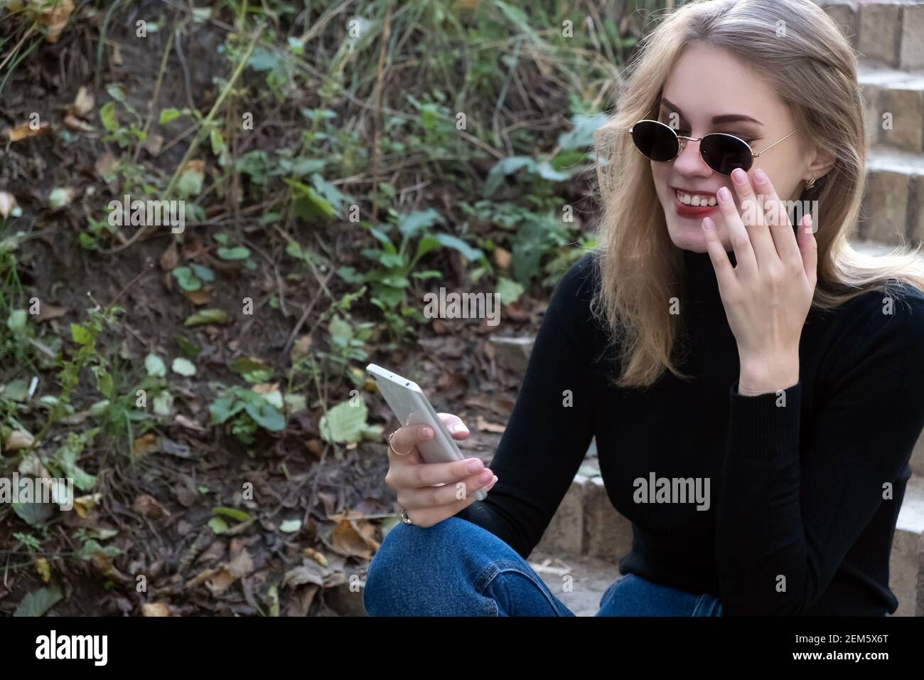 Young happy woman in black turtleneck looks into a mobile phone touching sunglasses with a hand while sitting on the steps in the park. Woman reads a Stock Photo