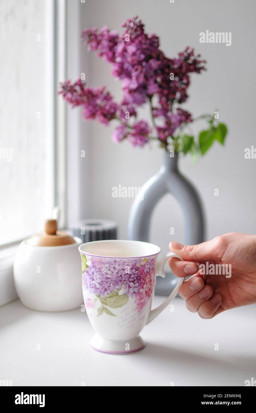 https://c8.alamy.com/comp/2EM694J/beautiful-composition-of-woman-hand-with-cup-of-tea-and-lilac-flowers-in-vase-2EM694J.jpg