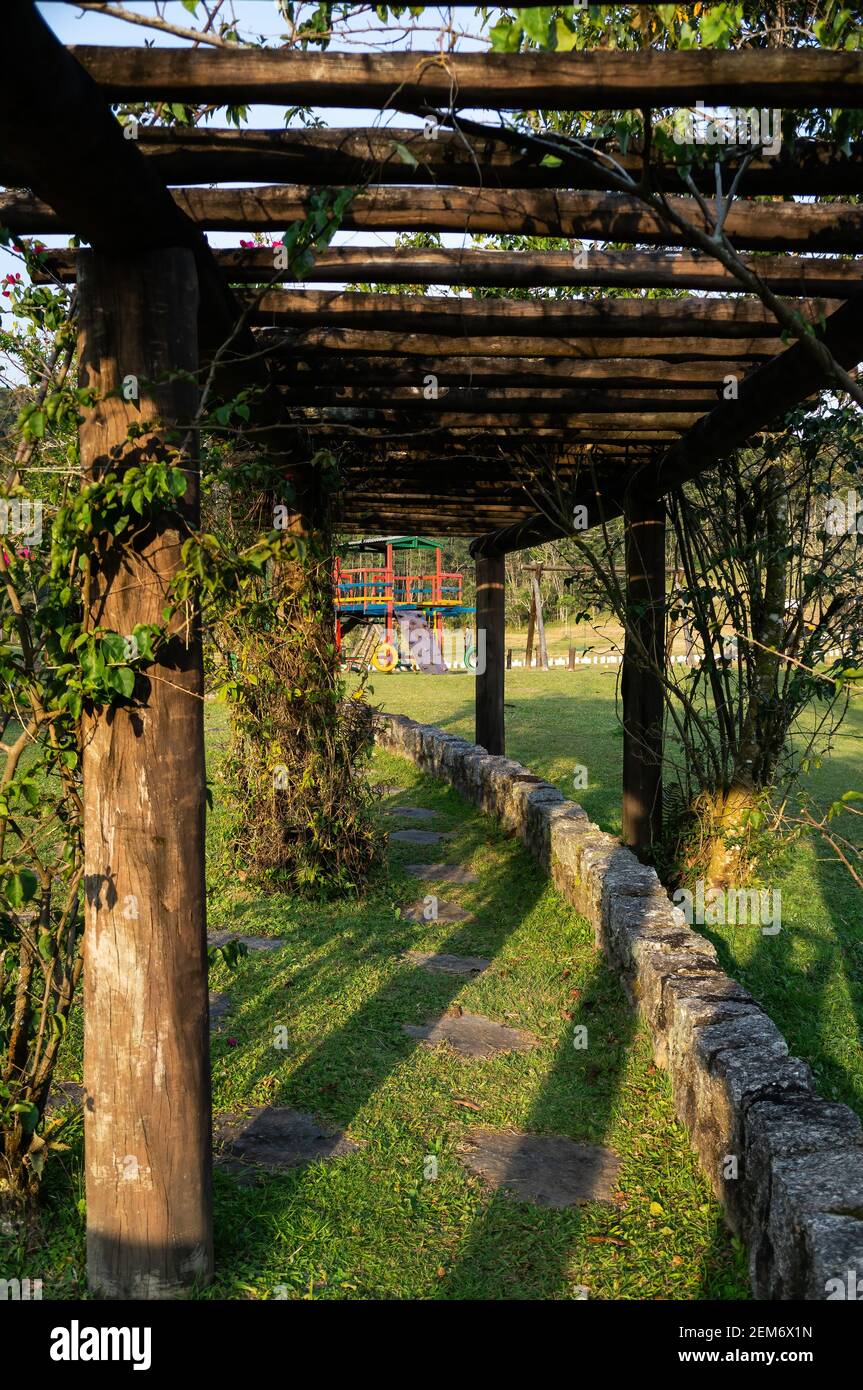 A curved narrow path under a wood structure for creeper plants near visitor's center and a playground of Serra do Mar estate park in late afternoon. Stock Photo