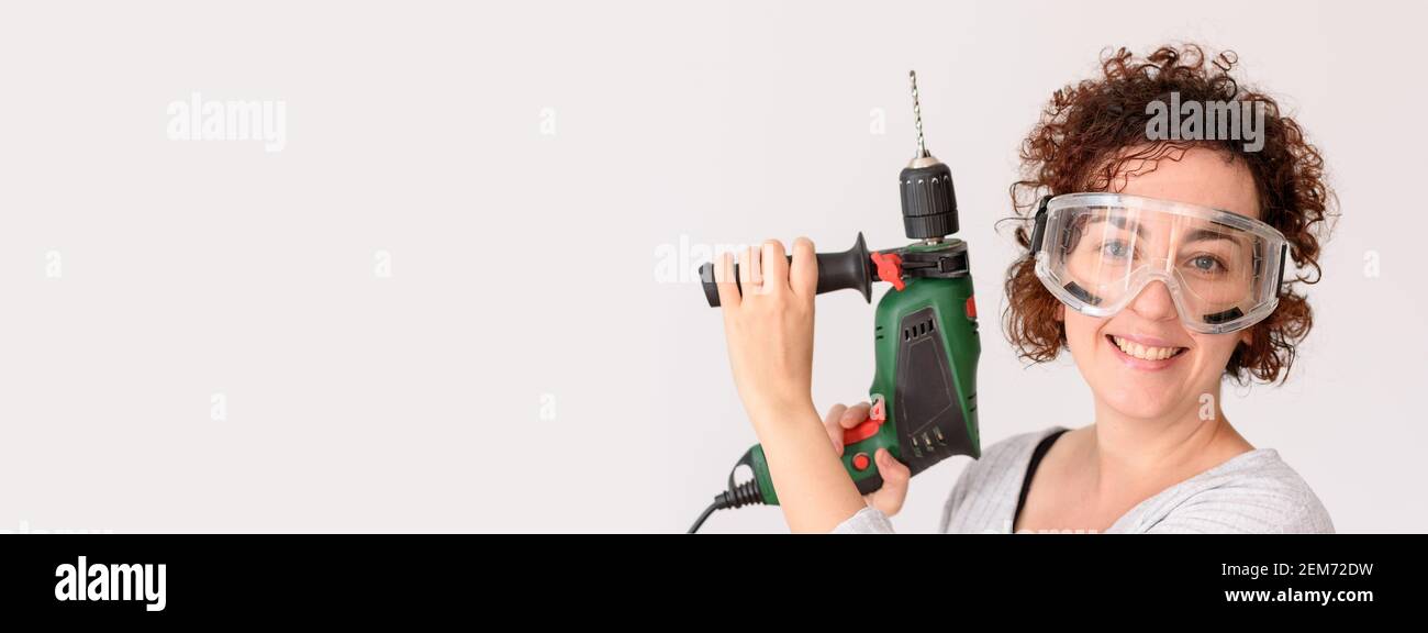 Caucasian woman with curly hair holds a drill in her hands, ready to make home improvements. She wears a gray shirt. White background. Natural light. Stock Photo