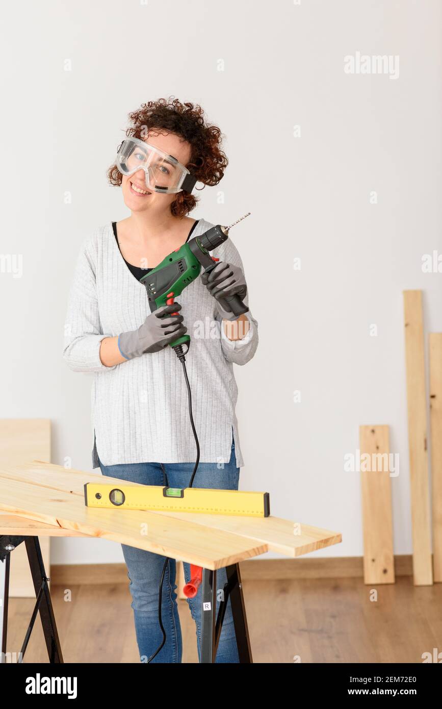 Caucasian woman with curly hair holds a drill in her hands, ready to make home improvements. She is standing in front of a workbench. She wears a gray Stock Photo