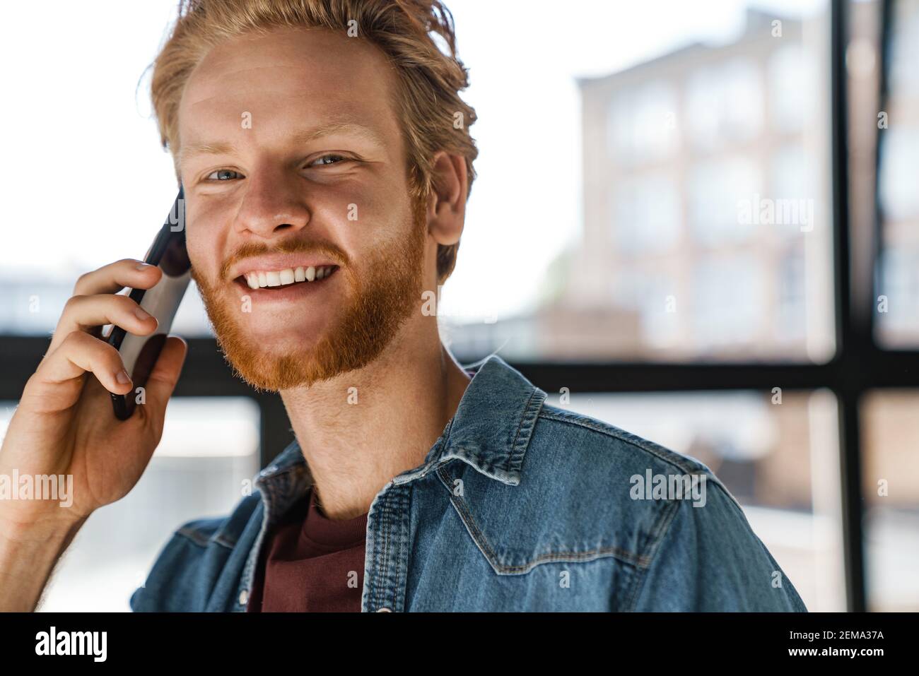 Happy handsome ginger guy smiling and talking on mobile phone indoors