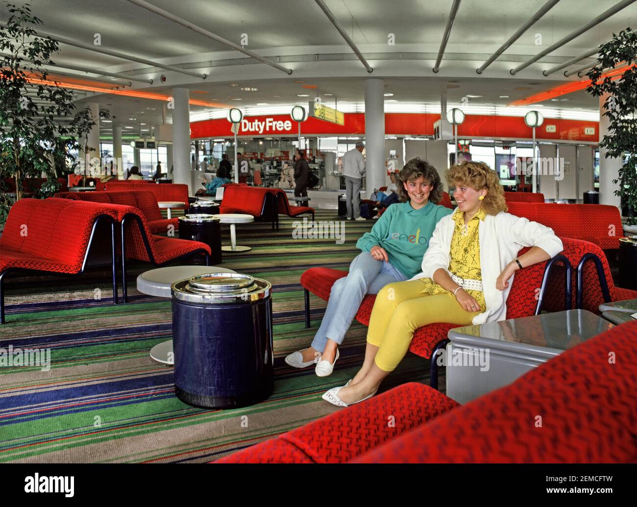 Two model released teenage girls talking and wearing 1980s fashion about to travel to 80s summer family holiday in Spain waiting in the departure lounge building with Duty Free shop & sign beyond at Gatwick Airport West Sussex England UK an historical archive image of the way we were in 1986 Stock Photo