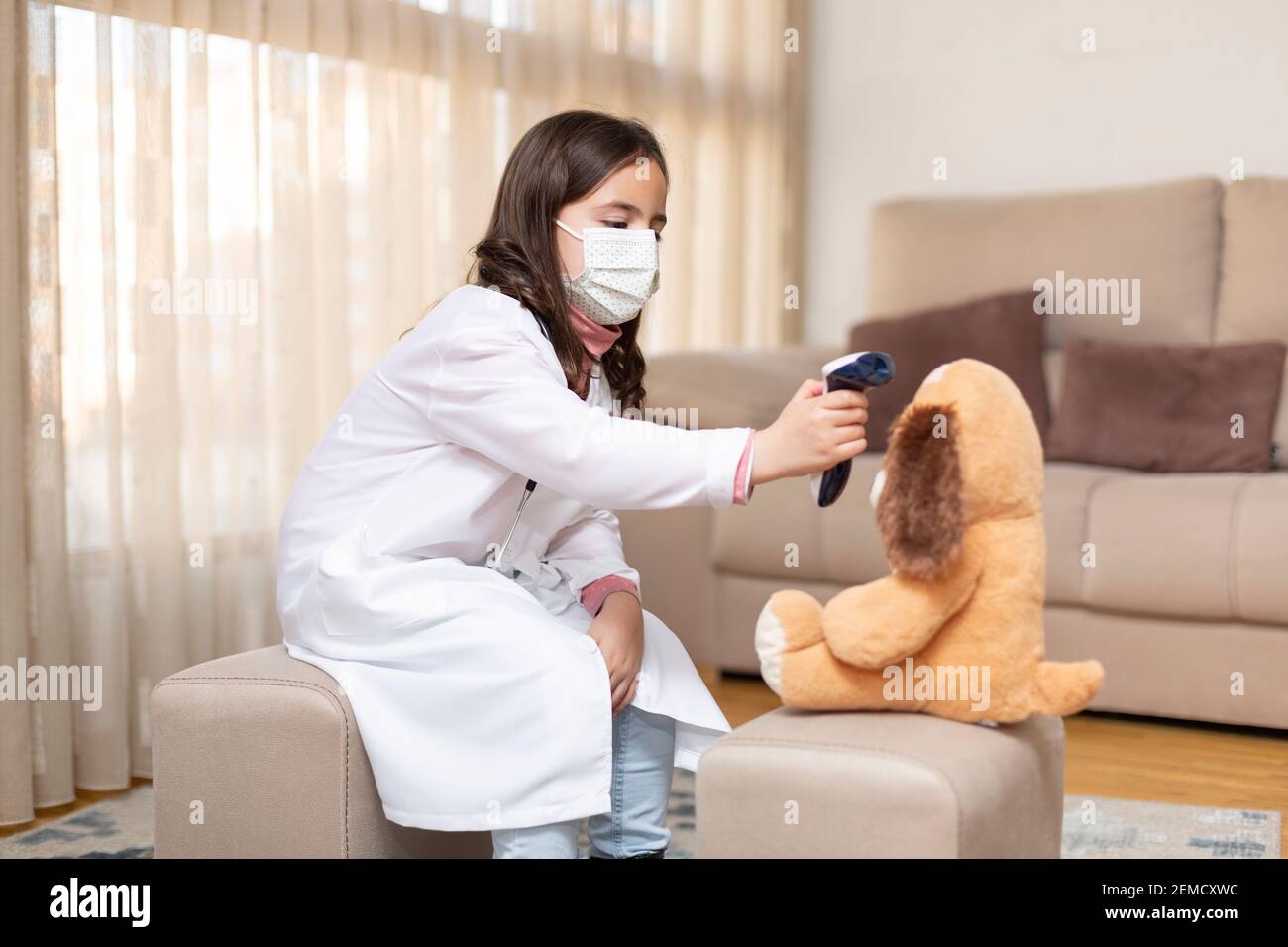 Little girl dressed as a doctor and medical mask taking the temperature of a teddy bear with an infrared thermometer. She is at home. Space for text. Stock Photo