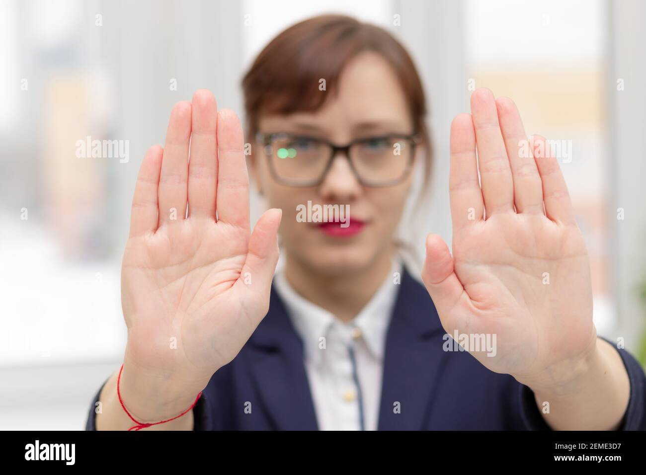 young strict business woman in a blue jacket and glasses at the window in the office at her workplace is unhappy and put her hands in front of her Stock Photo