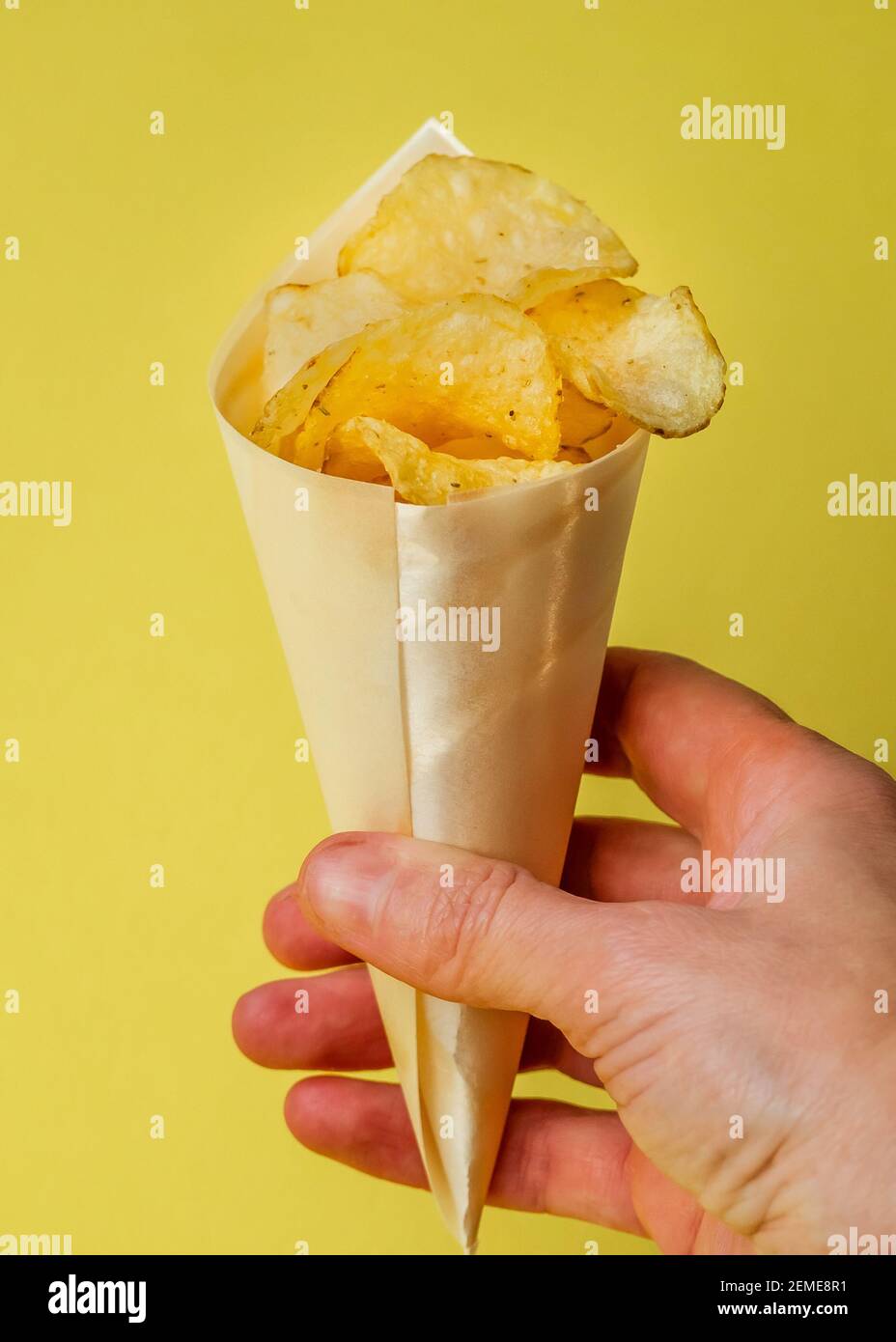 food yellow background. Salted potatoes chips. Hand holding a cone filled of golden fried chips on yellow. Fast salty food for snack or party. Tasty f Stock Photo