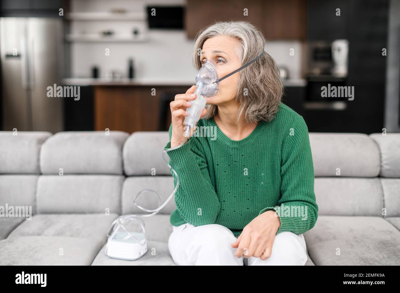 Sick senior woman has a viral infection or flu, labored breathing, asthma.  An elderly female is using inhaler, nebulizer for self treatment sitting  alone on the sofa at home Stock Photo -