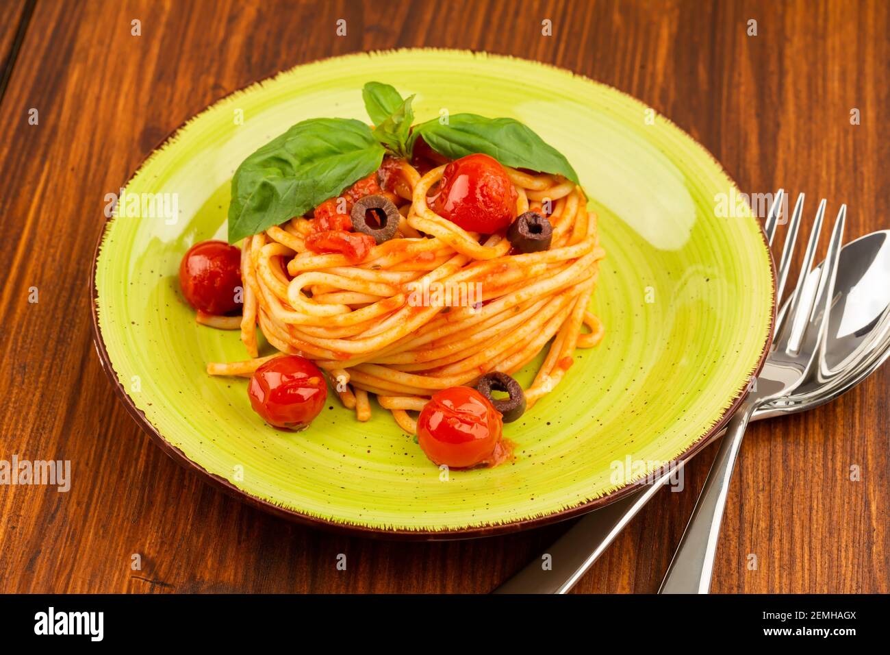 Pasta spaghetti Napoli or Napolitana on green plate with fork and spoon on brown background. Italian cuisine. Stock Photo