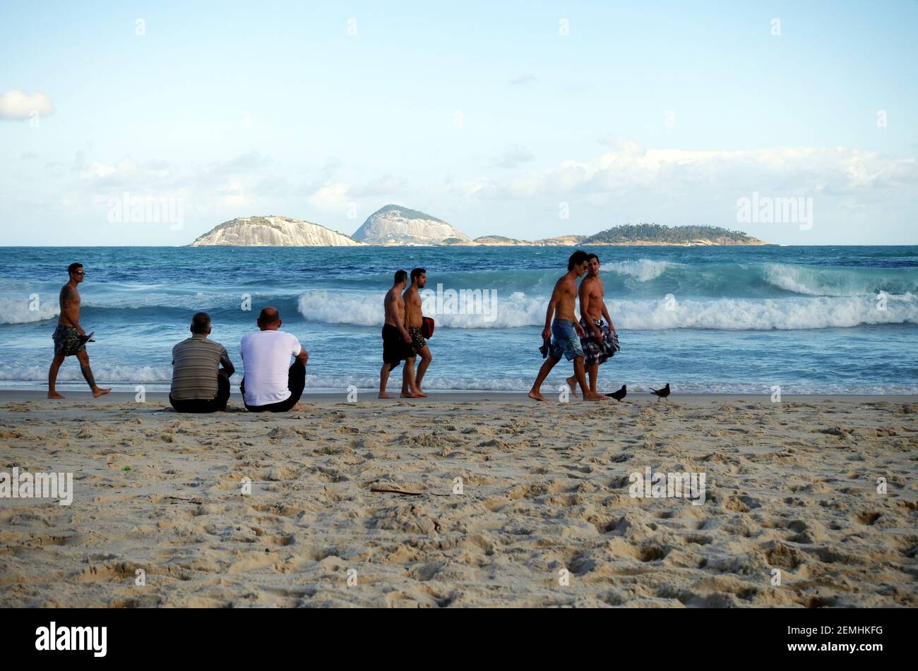 Two men sitting on Copacabana Beach, Rio de Janeiro, whilst some younger men stroll by along the waters edge Stock Photo
