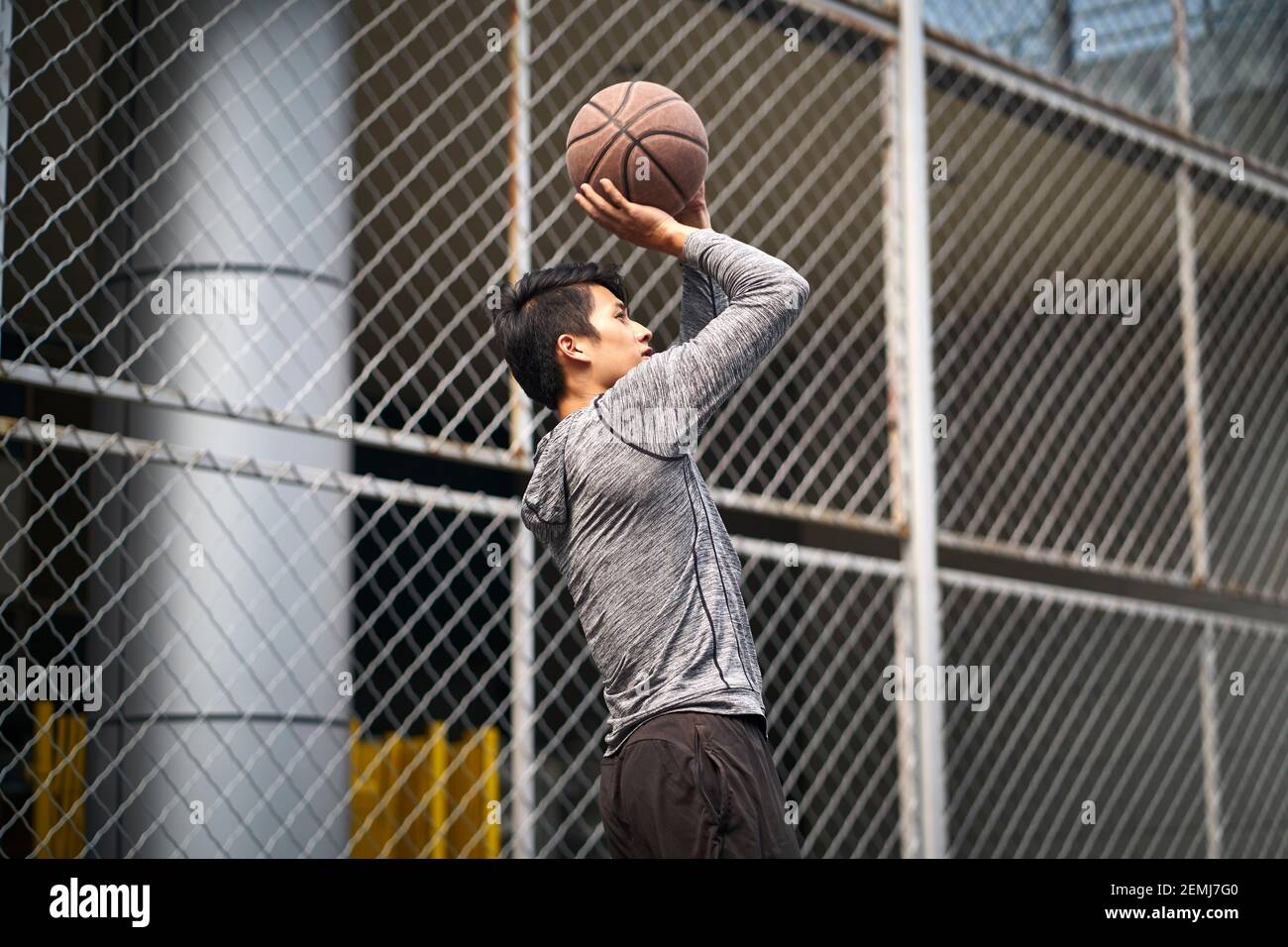 young asian male basketball player taking a jump shot on a fenced outdoor court Stock Photo