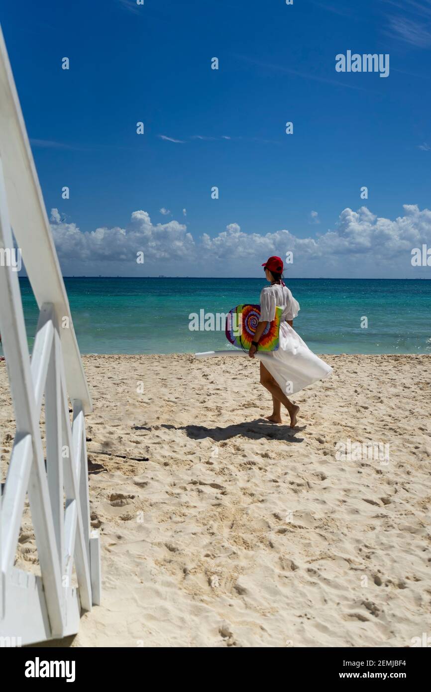 An attractive woman walks on a tropical beach with a white dress and a surfboard in Mexico. Caribbean sea and blue sky in the background. Concept travel Stock Photo
