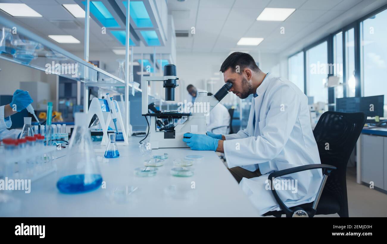 Modern Medical Research Laboratory: Male Scientist Working with Microscope, Analysing Biochemicals Samples. Advanced Scientific Lab for Medicine Stock Photo