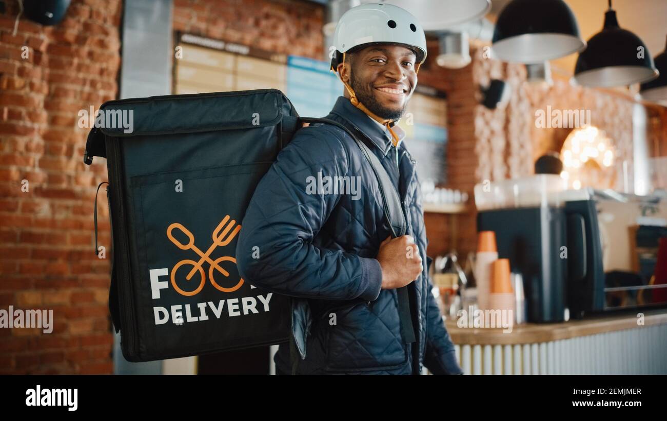 Handsome Black African American Food Delivery Courier Posing in Front of the Camera in a Coffee Shop. Happy and Smiling Man Wearing a Bicycle Helmet Stock Photo