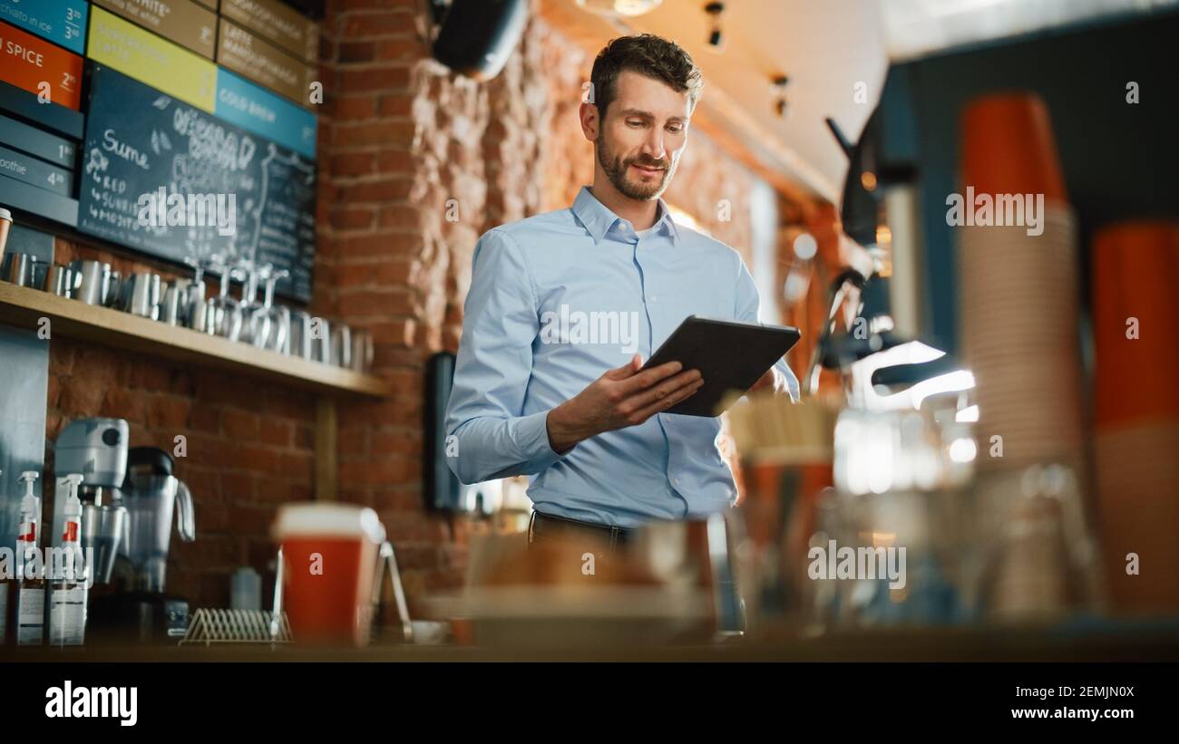 Handsome Caucasian Coffee Shop Owner is Working on Tablet Computer and Checking Inventory in a Cozy Loft-Style Cafe. Successful Restaurant Manager Stock Photo