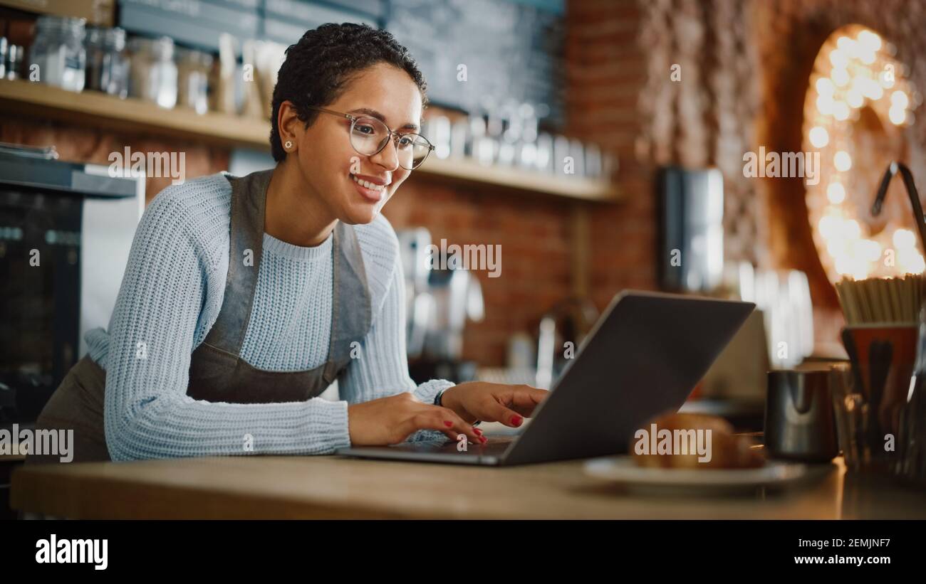 Young and Beautiful Latina Coffee Shop Owner is Working on Laptop Computer and Checking Inventory in a Cozy Cafe. Successful Restaurant Manager Stock Photo