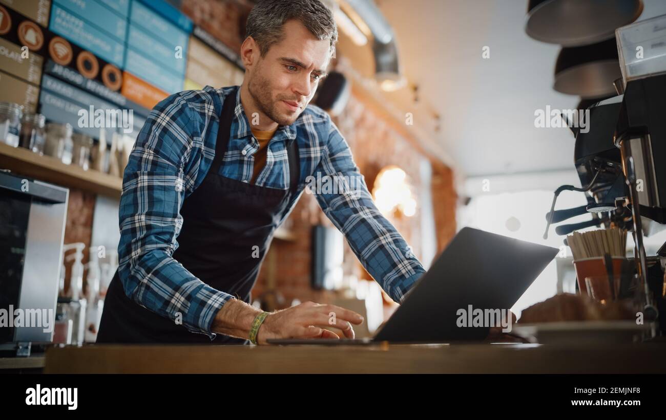 Young and Handsome Coffee Shop Owner is Working on Laptop Computer and Checking Inventory in a Cozy Cafe. Happy Restaurant Manager in Checkered Shirt Stock Photo