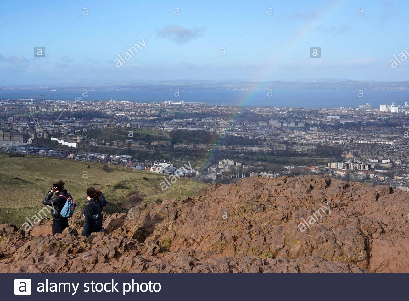 Edinburgh, Scotland, UK. 25th Feb 2021. People enjoying the sun and outdoors in Holyrood Park, on the summit of Arthurs Seat a rainbow appears after a rain shower.  Credit: Craig Brown/Alamy Live News Stock Photo