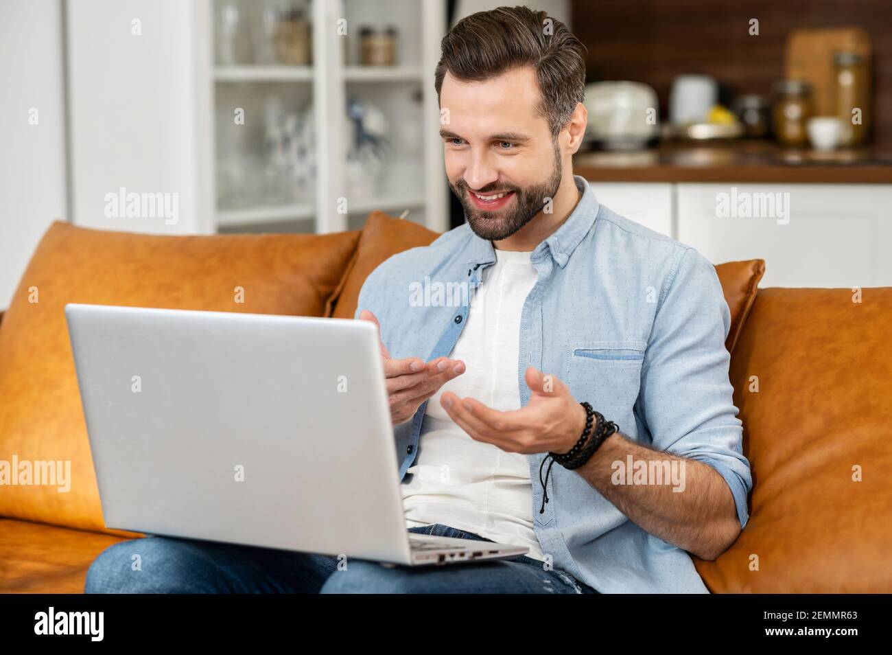 Smiling young bearded hipster guy tutor or a blogger sitting on the couch, streaming video online on the laptop, talking and explaining the strategy and gesturing, chatting in social media network Stock Photo
