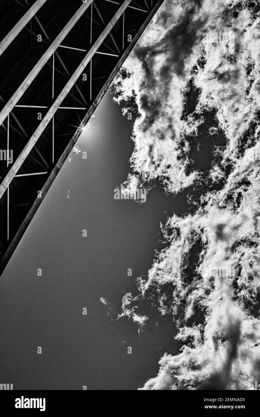 The lines of steel beams underneath the overpass create a striking contrast to the blue sky, clouds, and sun flare. Stock Photo