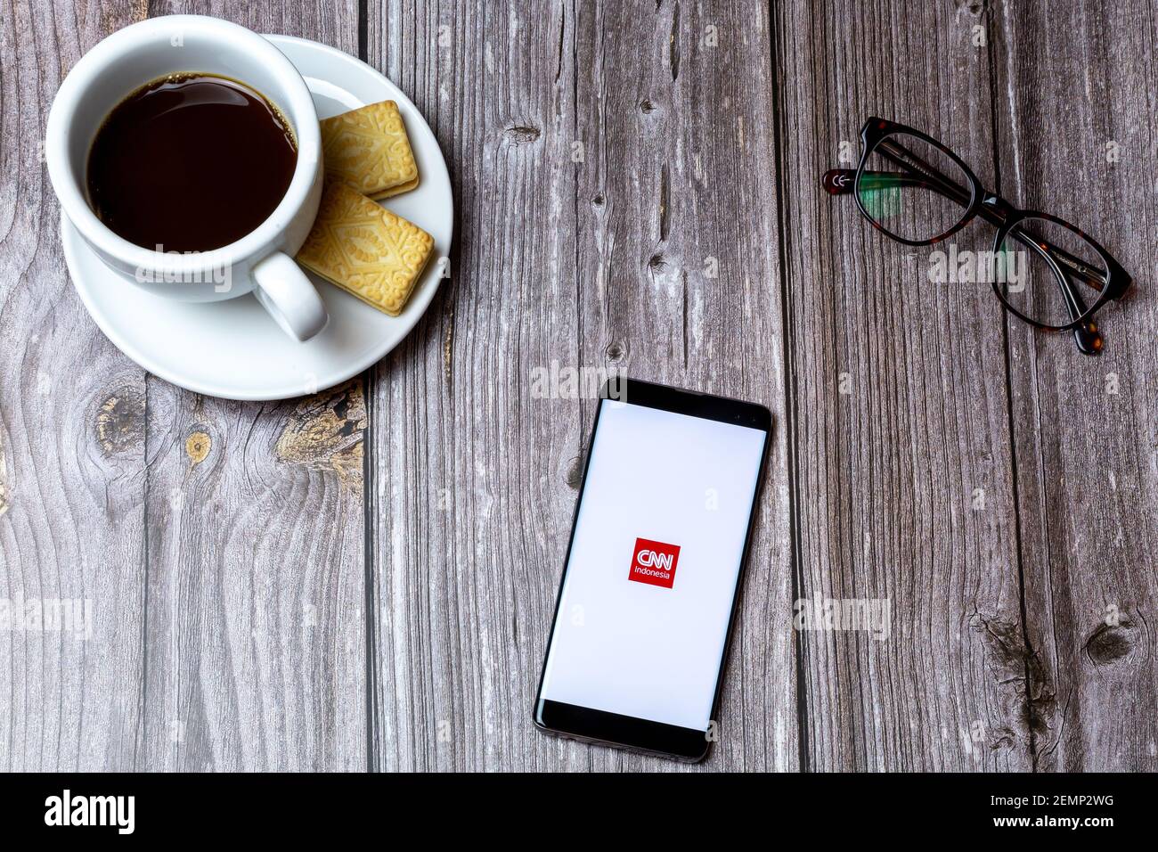 A mobile phone or cell phone on a wooden table with the CNN Indonesia app open next to a coffee and glasses Stock Photo