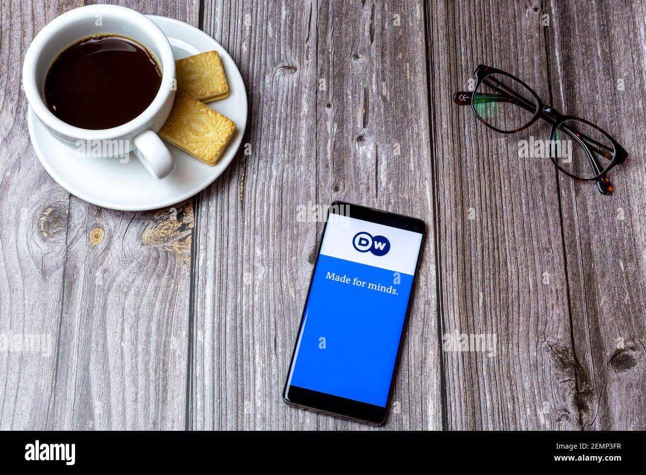 A mobile phone or cell phone on a wooden table with the Deutsche welle app open next to a coffee and glasses Stock Photo