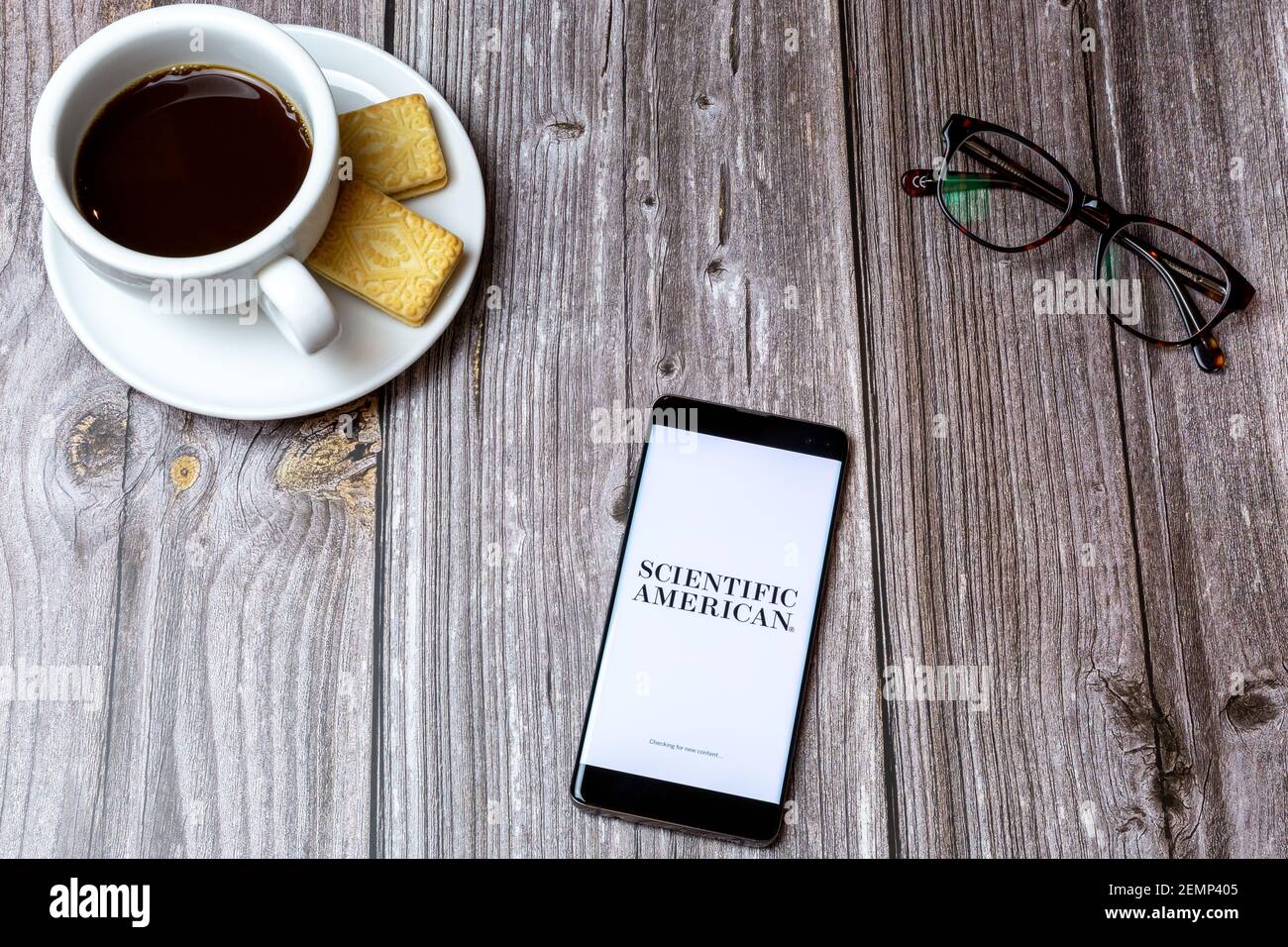 A mobile phone or cell phone on a wooden table with the Scientific American app open next to a coffee and glasses Stock Photo
