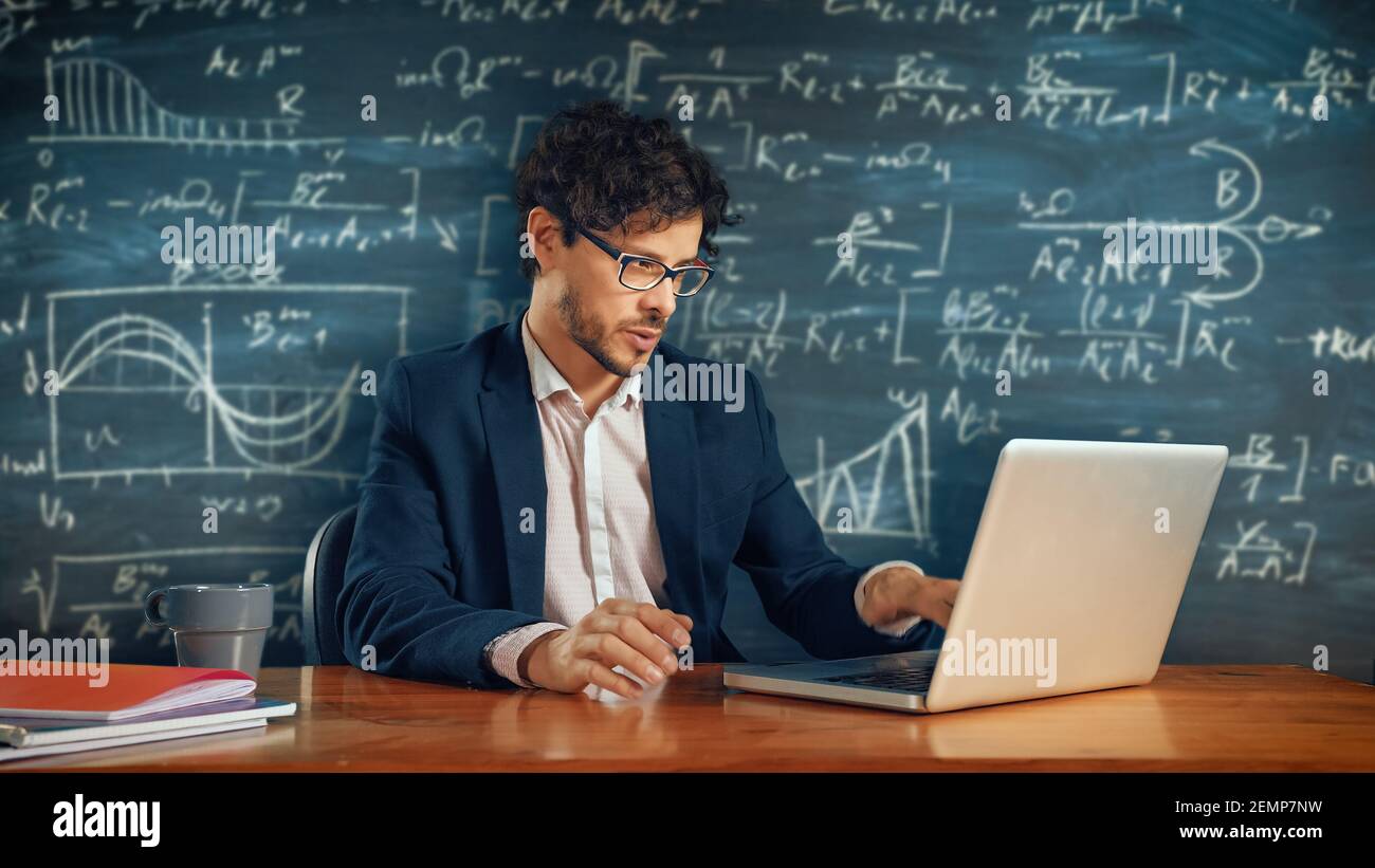 E-Education Concept: Teacher Sitting at Desk, Explains Lesson to a Classroom Using Laptop Computer, Blackboard Behind. e-Learning, Online Courses Stock Photo