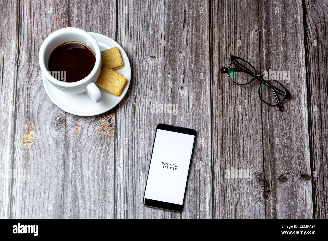 A mobile phone or cell phone on a wooden table with the Business Insider app open next to a coffee and glasses Stock Photo