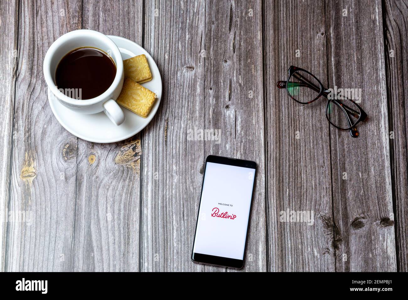 A mobile phone or cell phone on a wooden table with the Butlins holidays app open next to a coffee and glasses Stock Photo