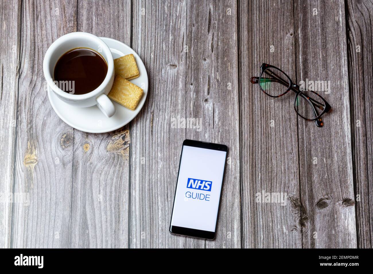 A mobile phone or cell phone on a wooden table with the NHS Guide app open next to a coffee and glasses Stock Photo