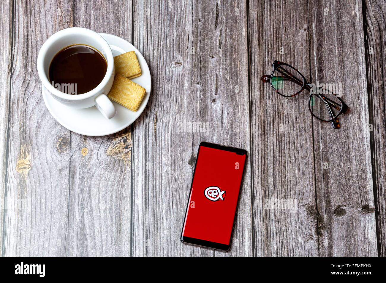 A mobile phone or cell phone on a wooden table with the CEX app open next to a coffee and glasses Stock Photo