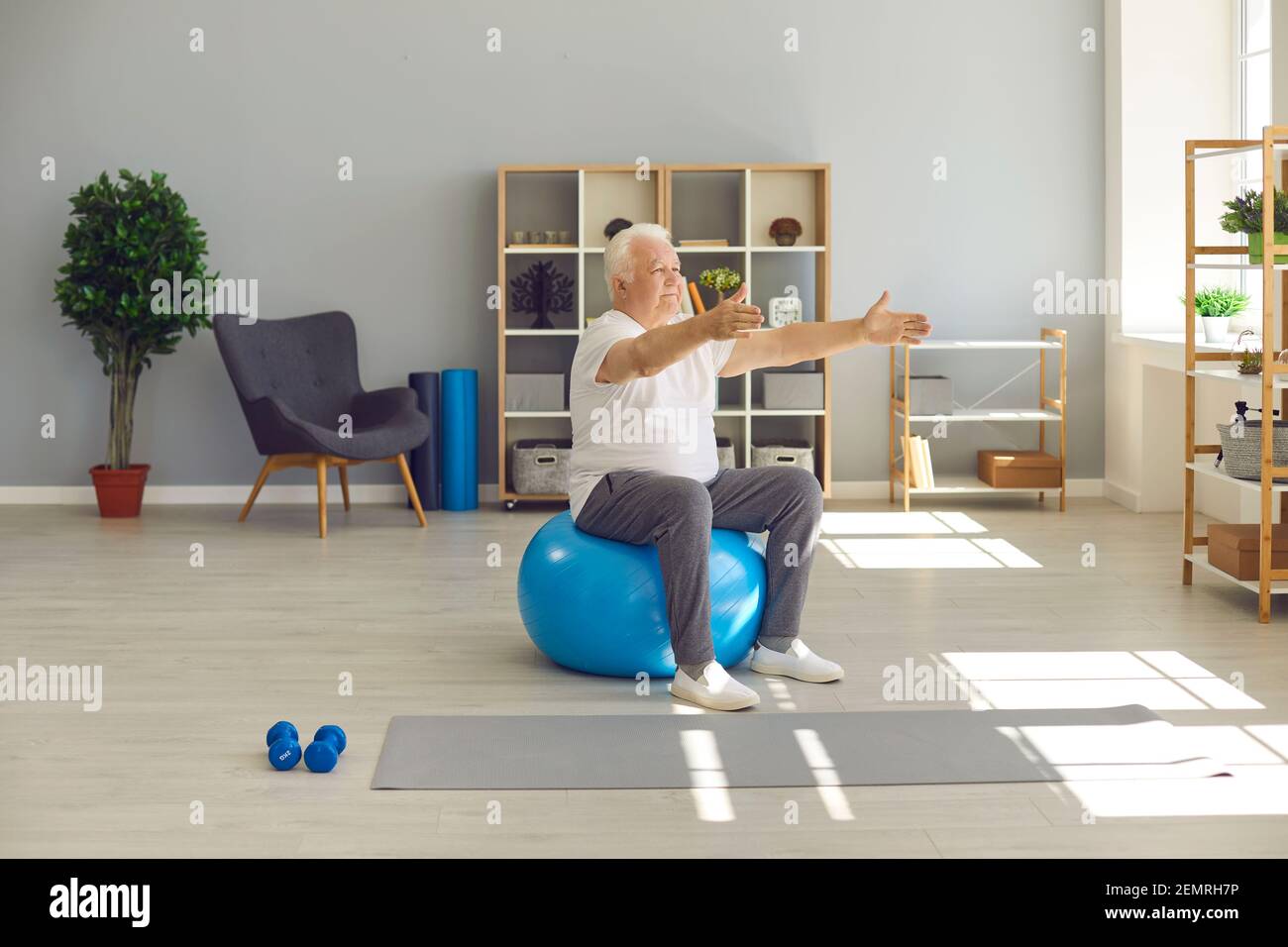 Elderly man sitting and exercising on fitness ball with arms stretched at home or in rehabilitation clinic Stock Photo
