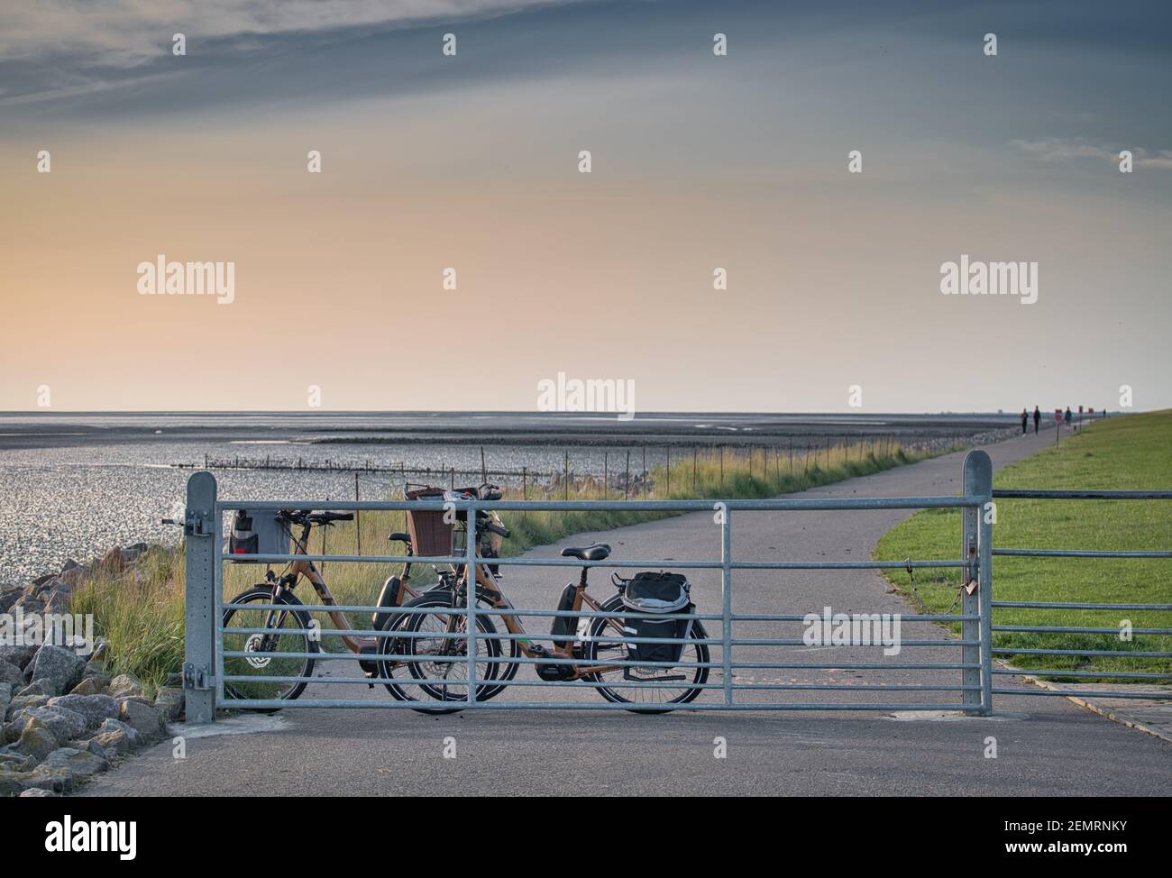 Bicycles leaning against a fence on the dike before sunset. A path leads along the sea over the dike. People are running on the horizon. Stock Photo