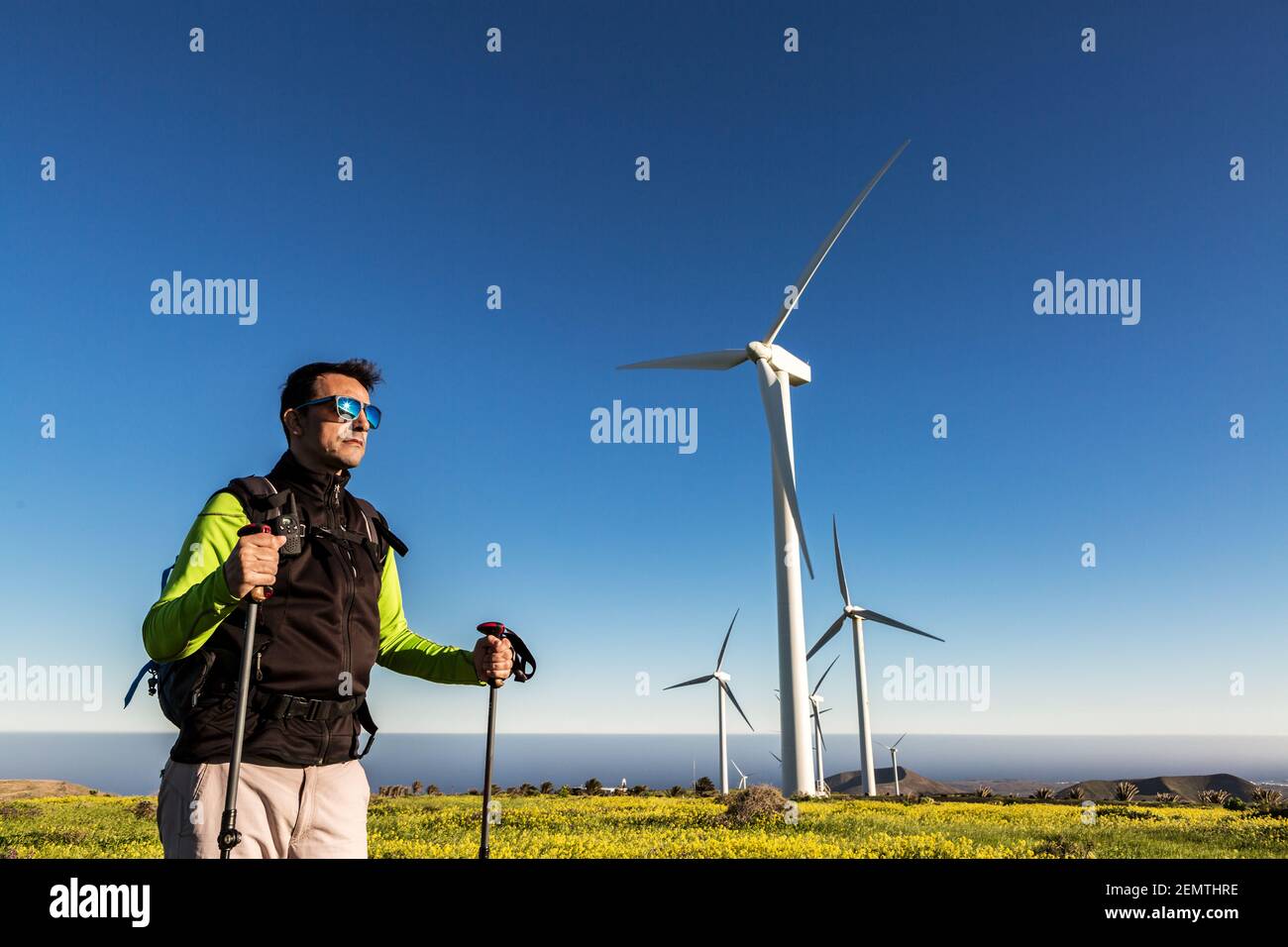 Male explorer with backpack and trekking poles standing in meadow with windmills on background of blue sky on Lanzarote and looking away Stock Photo