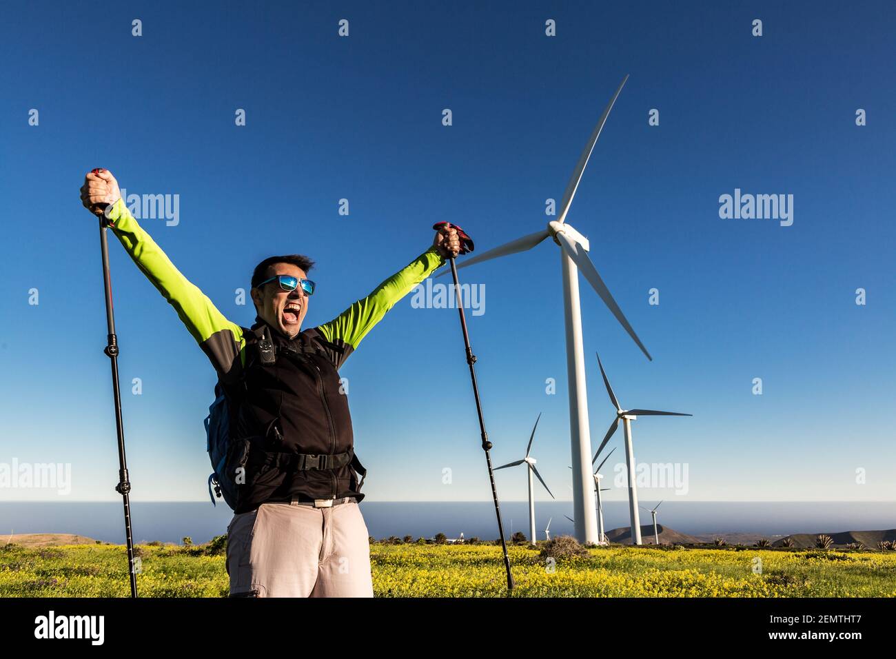 Expressive male hiker with trekking poles standing with raised arms and celebrating goal achievement on background of windmills on Lanzarote Stock Photo