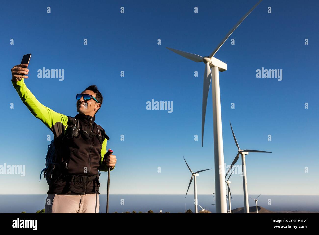 Cheerful male adventurer with trekking poles and backpack taking selfie on smartphone while standing on background of windmills on Lanzarote Stock Photo