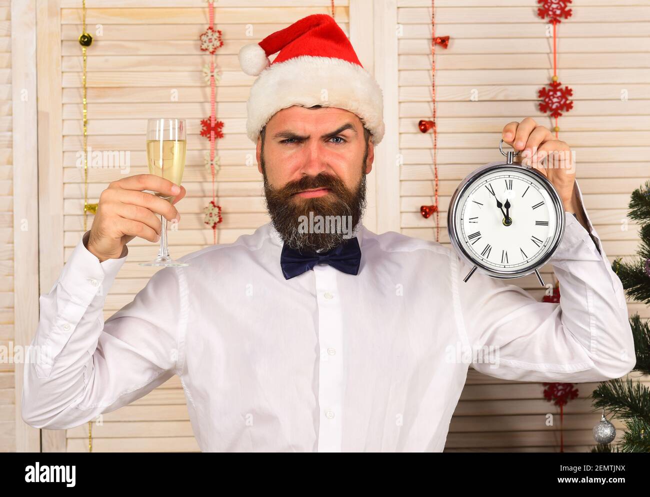 Santa Claus in red hat with angry face in festive room. Guy on wooden wall background with garlands. Celebration and New Year time concept. Man with beard holds glass of champagne and alarm clock Stock Photo