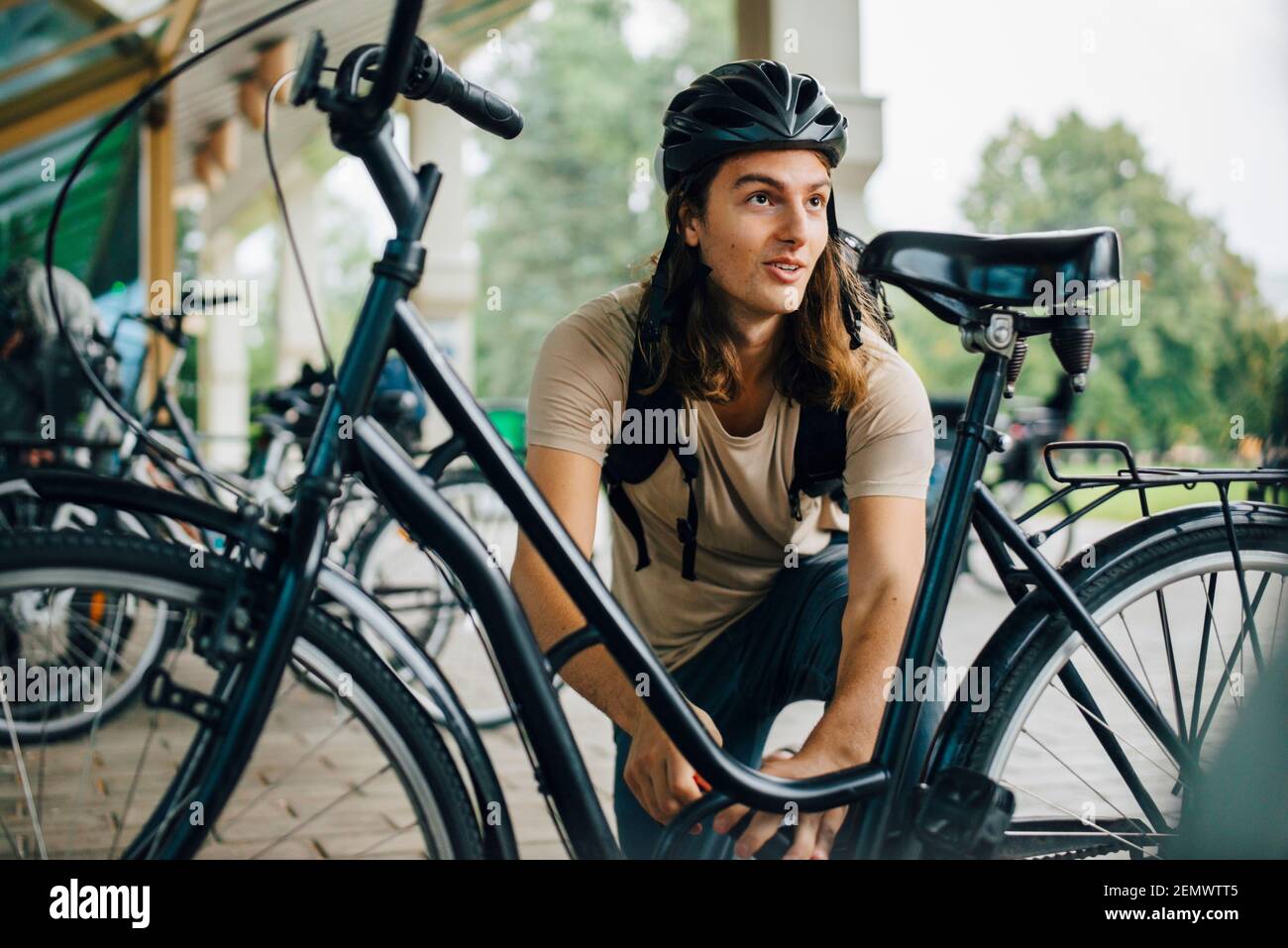 Male student looking away while unlocking bicycle at parking station in college campus Stock Photo