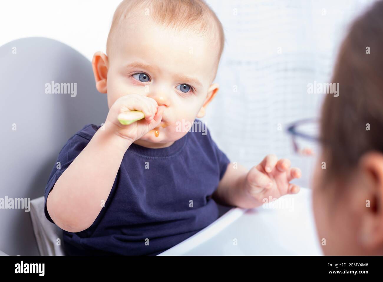Cute caucasian baby boy is sitting in high chair and eating with plastic spoon. Mom is proud of her son, doesnt help him. Baby is eating on his own. Stock Photo