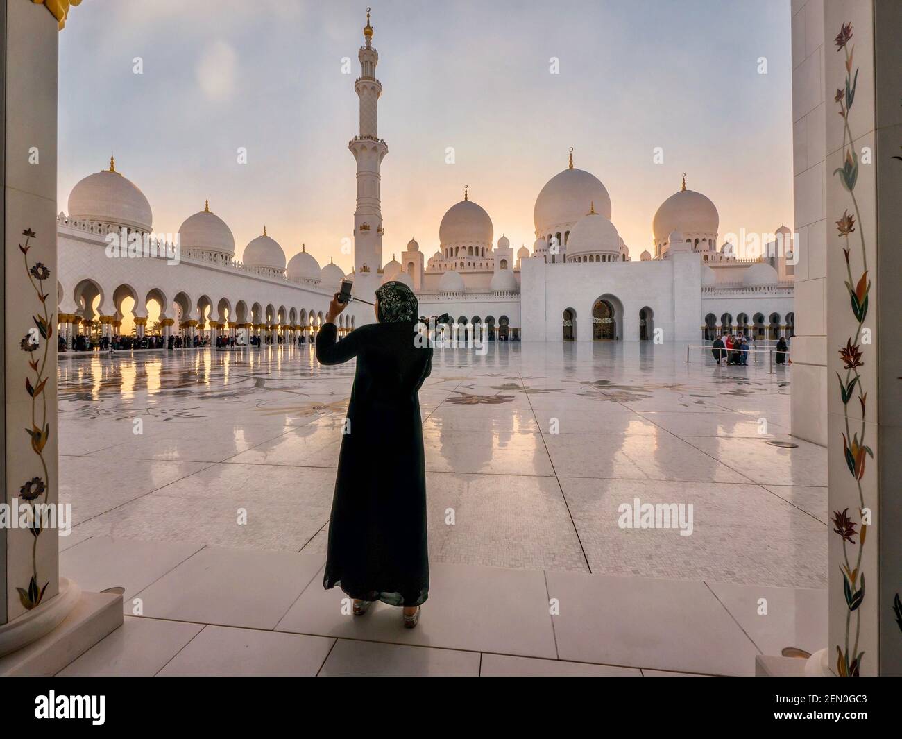A young Muslim woman wearing a black abaya takes a selfie at a very beautiful mosque at sunset in Abu Dhabi, UAE. Stock Photo