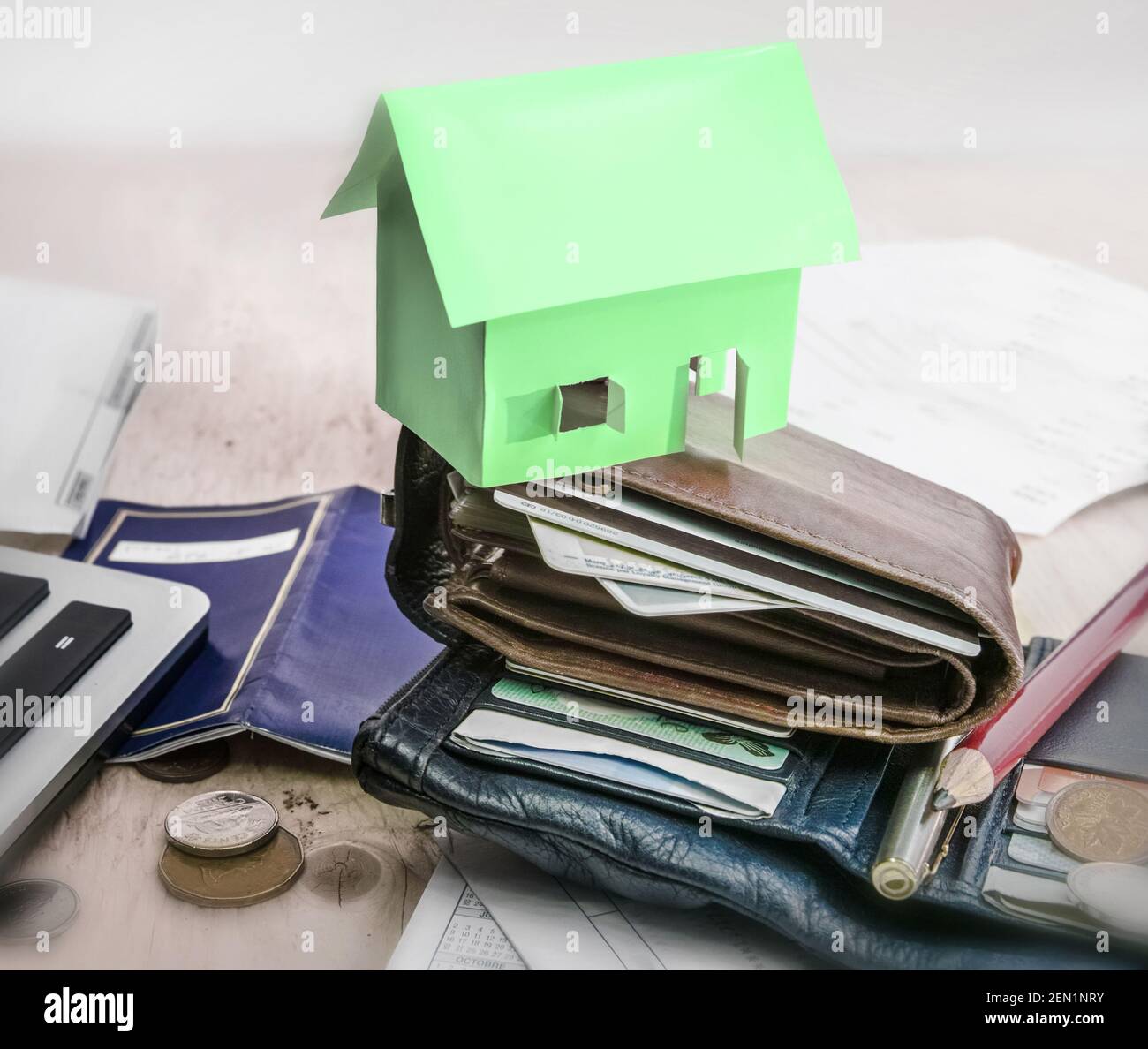 Above a stack of wallets sits a small green paper house. On the table, all around, coins, various papers, pencil and pen Stock Photo