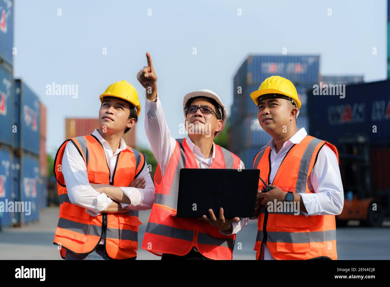 Asian foreman holding laptop and explaining the various operations in the container depot terminal to worker. Stock Photo