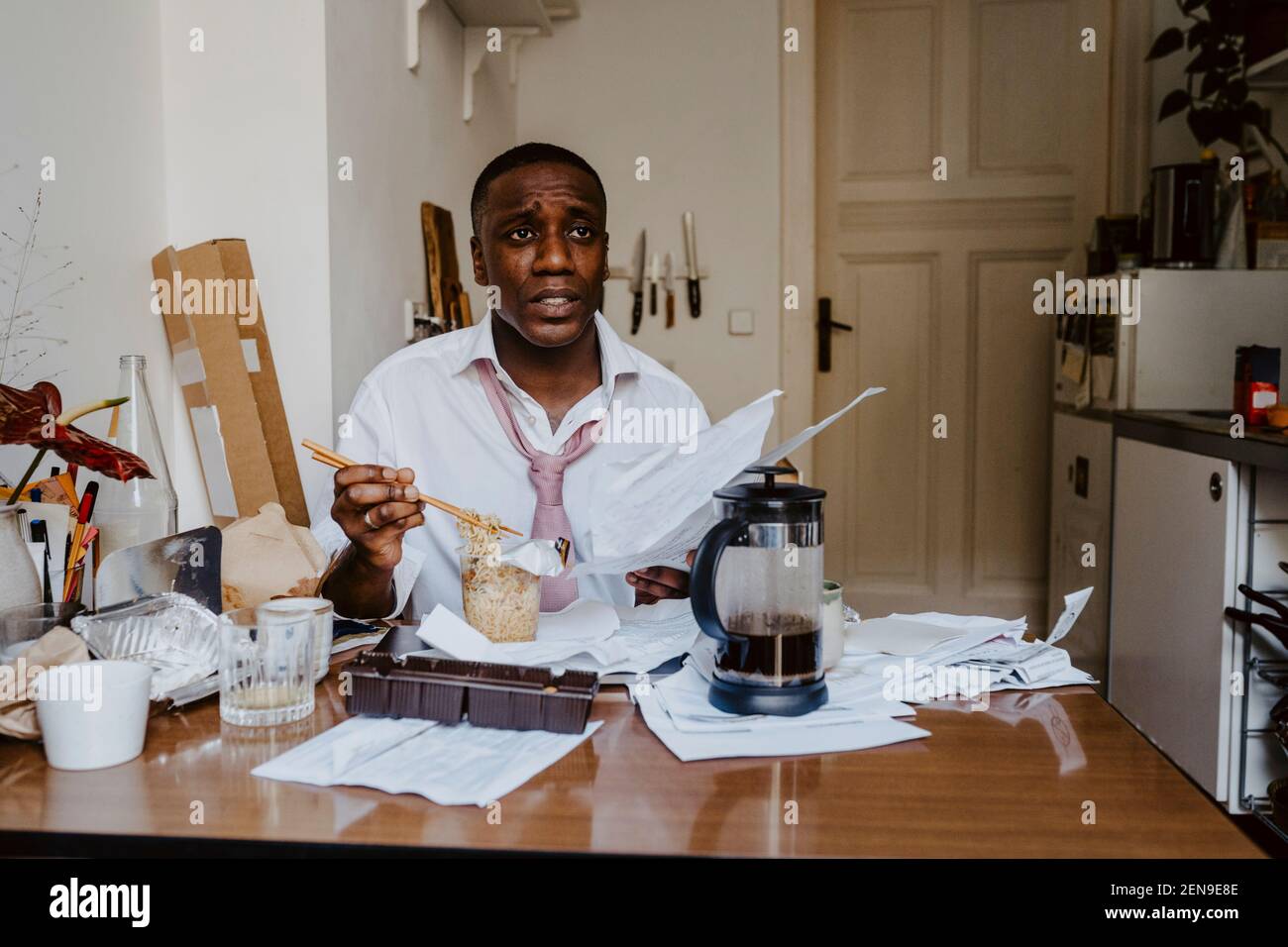 Thoughtful male entrepreneur with paper document having noodles at table in living room Stock Photo