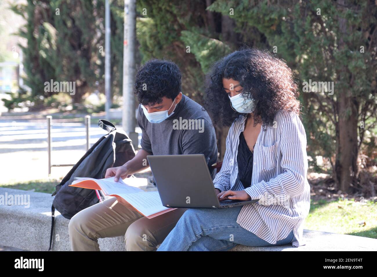 Two latin students wearing protective face mask studying together sitting on a bench outdoor. New normal at university campus. Stock Photo