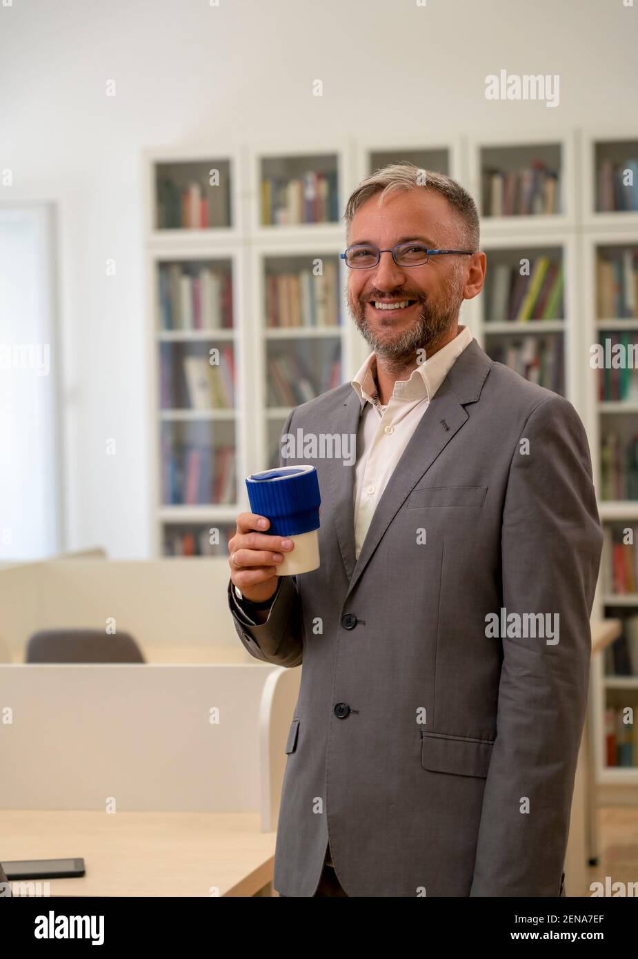 Happy Middle Aged Businessman Standing at Office Desk During Coffee Break. Portrait of Mature Man With Cup of Coffee. Stock Photo