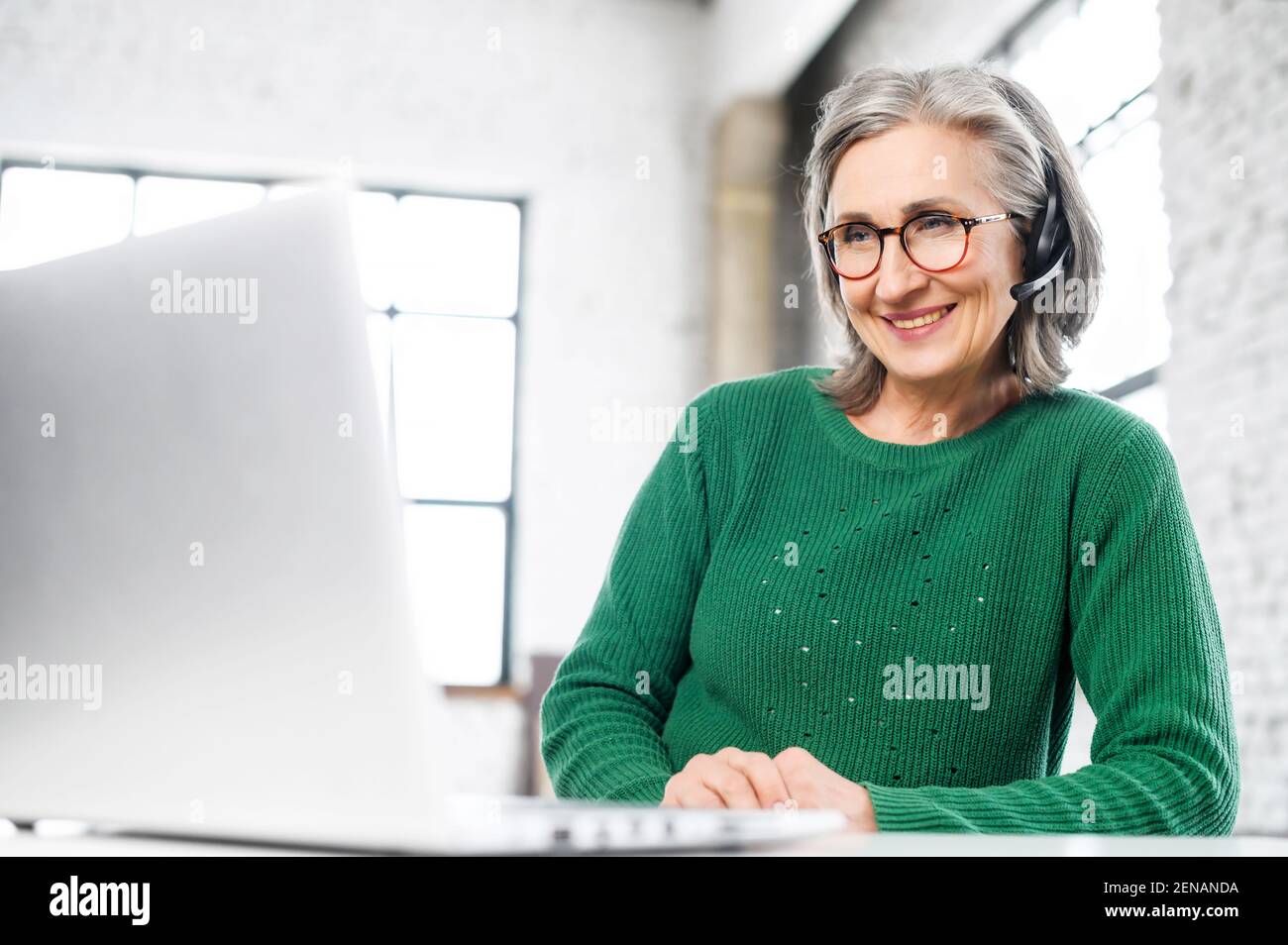 Cheerful aged woman with headset, wrinkles on the face, smiling, listening at the computer, consulting the client online , gives the advice, home working, positive person, takes every chance to work Stock Photo