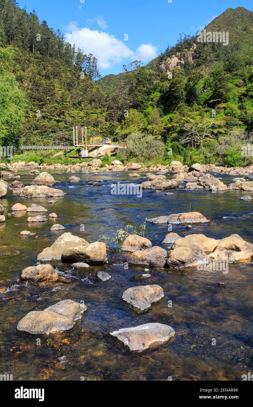 The rocky Ohinemuri River flowing through the Karangahake Gorge, New Zealand. A suspension bridge crosses the river in the background Stock Photo