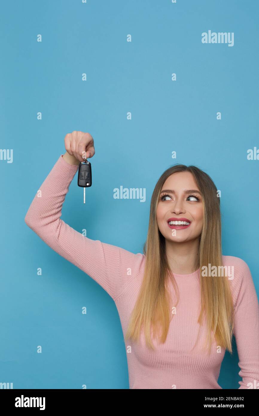 Happy young woman in pink sweater is holding car key and looking at it. Waist up studio shot on blue background. Stock Photo