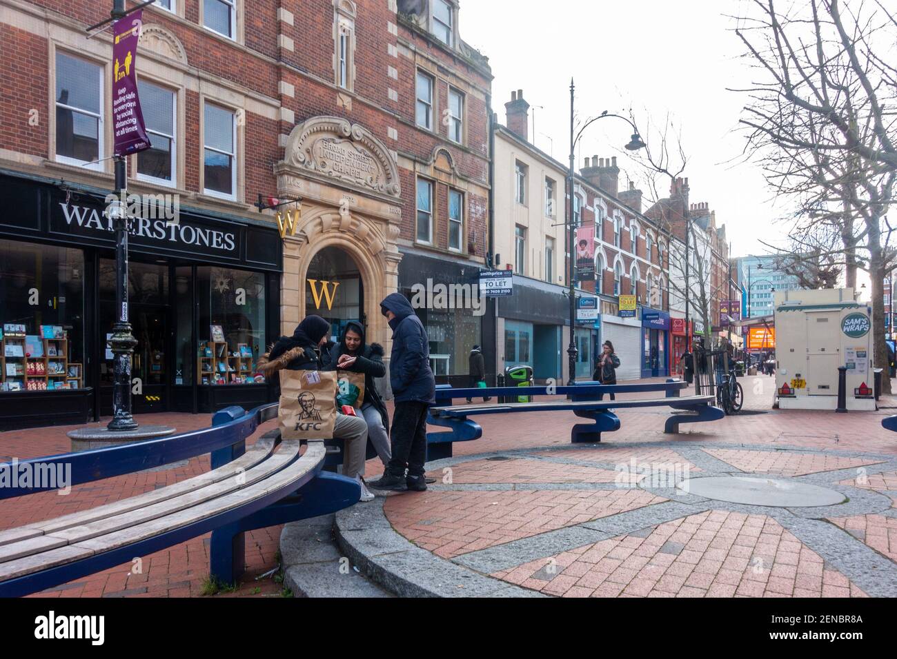 Three girls sit on a bench together in what is an otherwise quiet Broad Street in Reading town centre in Berkshire UK.. Shops are shut due to COVID-19 Stock Photo