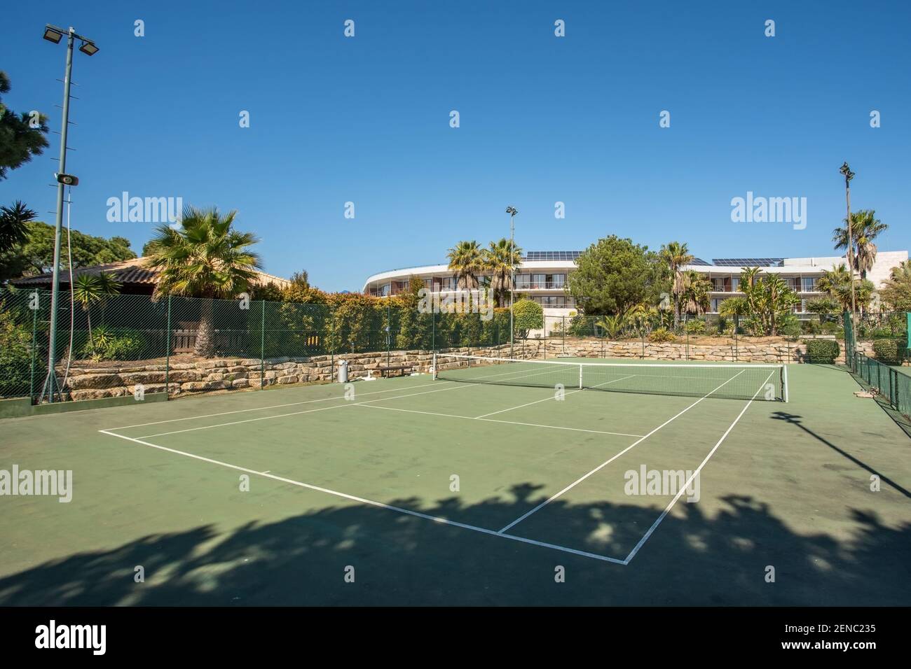 Tennis court seen from the corner, surrounded by lots of green trees and a building a bit further in front, on a bright summer day. Stock Photo